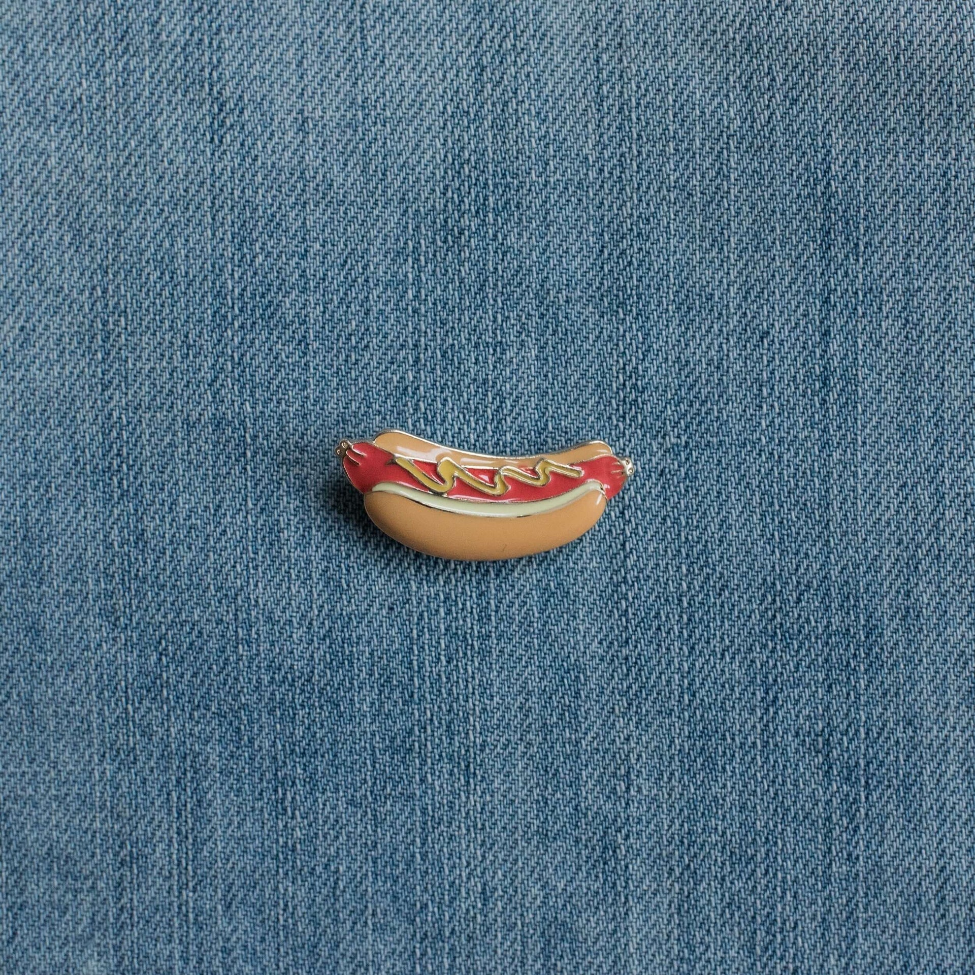 Hot dog enamel pin with ketchup and mustard on denim fabric background