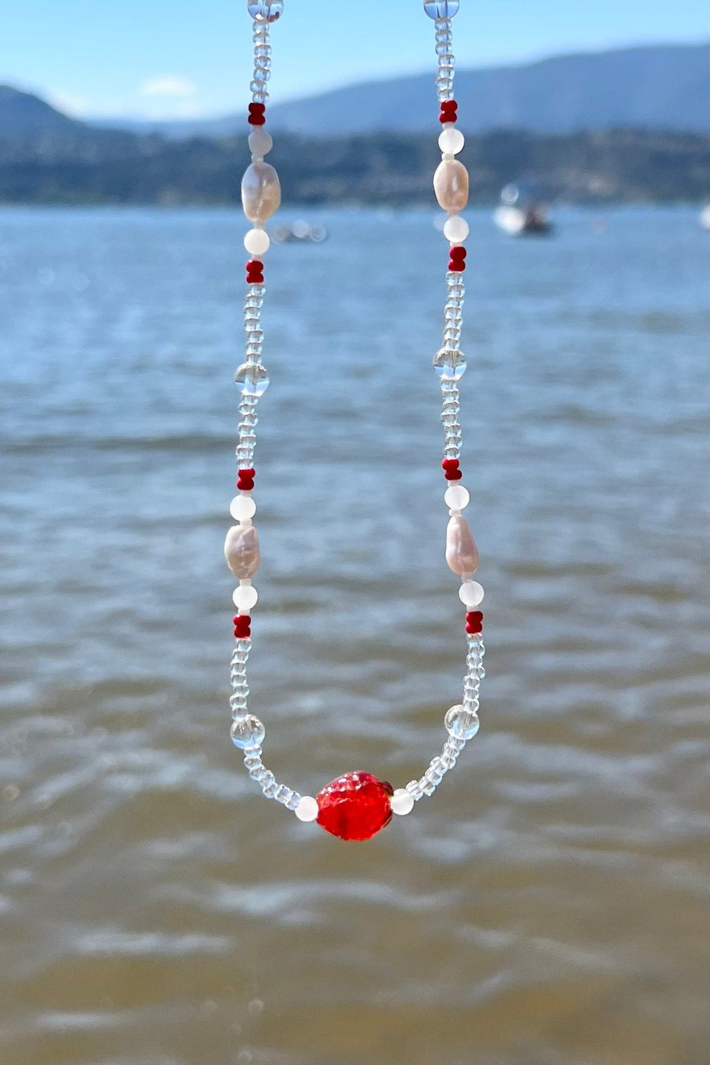 Handmade beaded necklace with red, clear, and white beads by the lake
